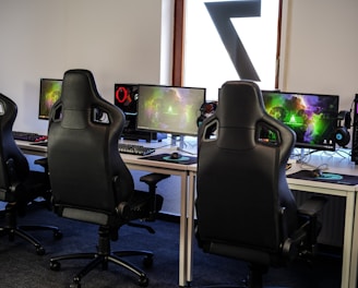 A row of gaming stations equipped with large monitors, gaming chairs, keyboards, and headphones. The setup is organized and ready for gaming, featuring a colorful screen display. The room has a modern and tech-savvy ambiance.