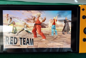 A handheld gaming console displays a screenshot of a fighting game featuring cartoonish characters in dynamic poses. The characters are standing in a virtual arena setting with a sky and cloud backdrop. The console is placed on a green surface.
