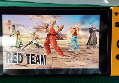 A handheld gaming console displays a screenshot of a fighting game featuring cartoonish characters in dynamic poses. The characters are standing in a virtual arena setting with a sky and cloud backdrop. The console is placed on a green surface.