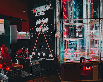 An arcade setting featuring a classic Space Invaders game setup and a modern gaming machine with seats. Bright neon lights illuminate the area, with scores displayed on digital screens. A bright and visually engaging environment full of electronic components.