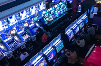 A bustling arcade filled with rows of gaming machines, each with vibrant displays and controls. Several people are seated at these machines, deeply engaged in playing games. Bright fluorescent lights cast a blue and neon glow across the room, enhancing the lively and energetic atmosphere.